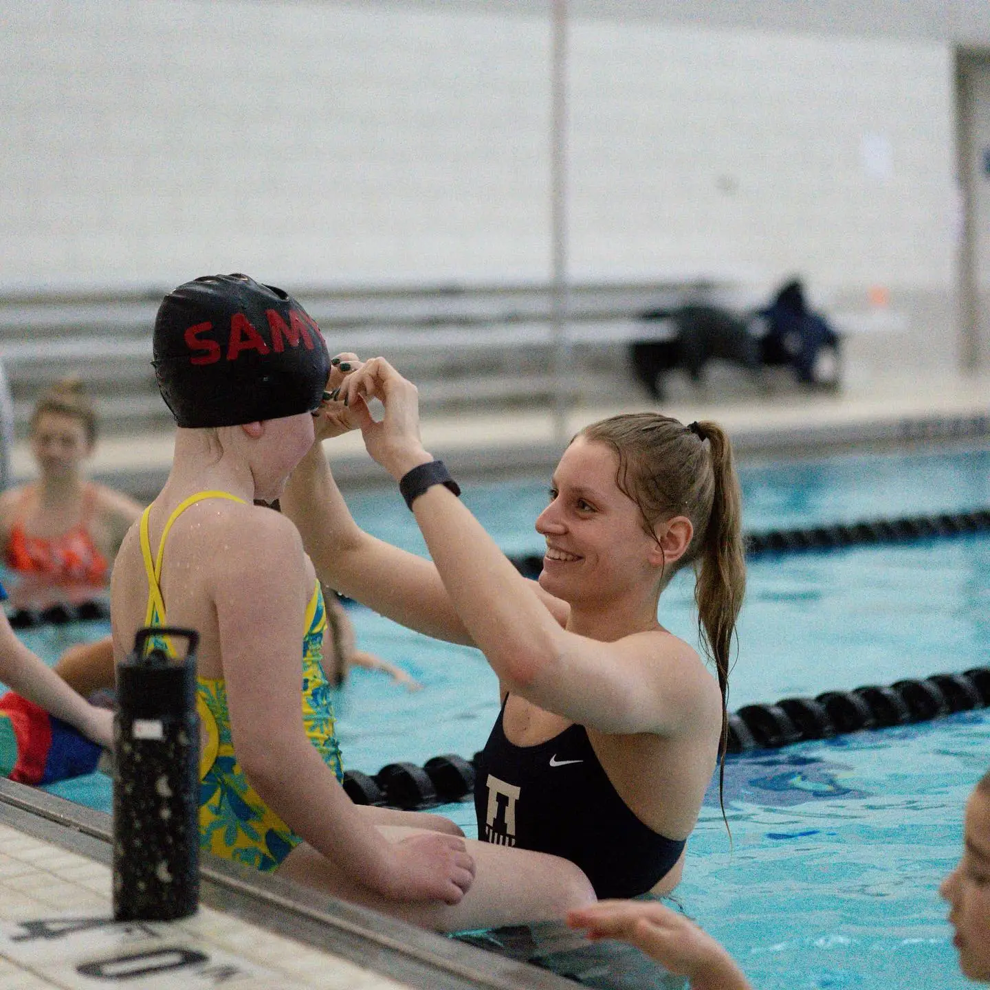 A college swimmer helping a young camper with her goggles poolside